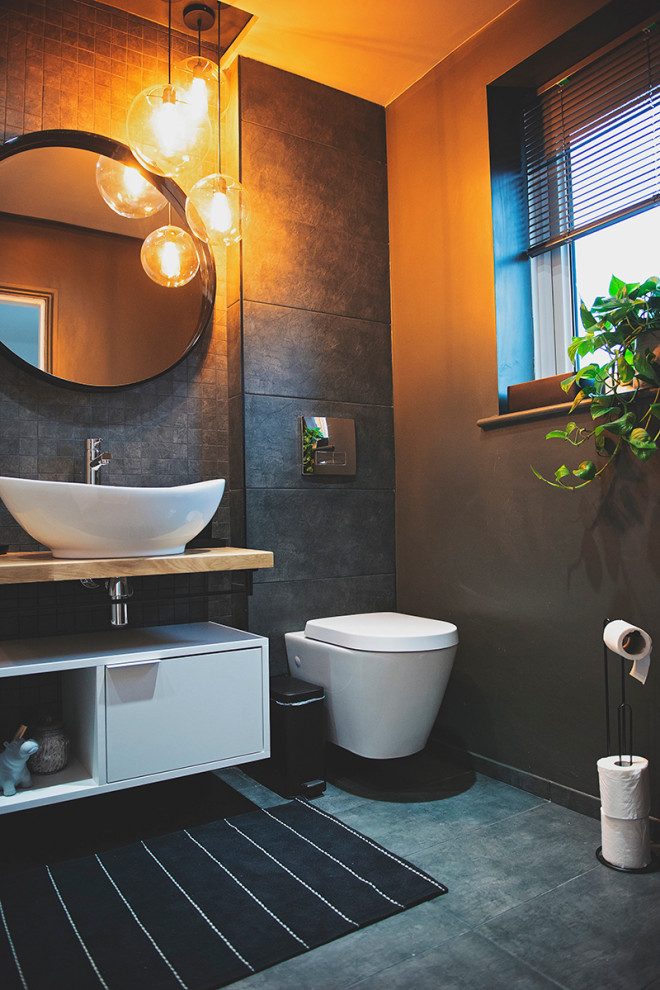 This is an example of a mid-sized modern bathroom in Manchester with furniture-like cabinets, white cabinets, an open shower, a wall-mount toilet, black tile, mosaic tile, grey walls, ceramic floors, a console sink, wood benchtops, black floor, an open shower, brown benchtops, a single vanity and a freestanding vanity.