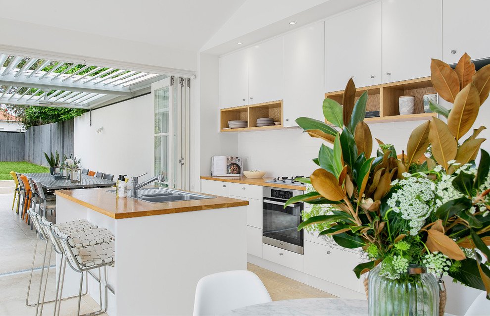This is an example of a small scandinavian galley eat-in kitchen in Sydney with a double-bowl sink, flat-panel cabinets, white cabinets, wood benchtops, white splashback, subway tile splashback, stainless steel appliances, travertine floors, with island and beige floor.