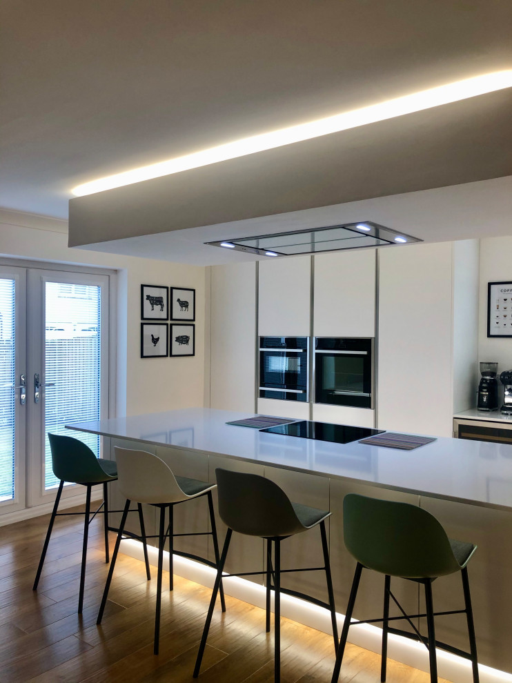 Photo of a large contemporary l-shaped eat-in kitchen in Glasgow with an integrated sink, flat-panel cabinets, white cabinets, quartzite benchtops, green splashback, glass sheet splashback, black appliances, vinyl floors, with island, brown floor, white benchtop and recessed.