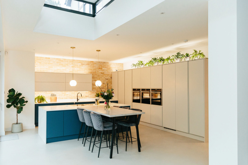 This is an example of a large traditional open plan kitchen in London with a built-in sink, flat-panel cabinets, blue cabinets, black appliances, cement flooring, an island, grey floors, white worktops, a vaulted ceiling and feature lighting.