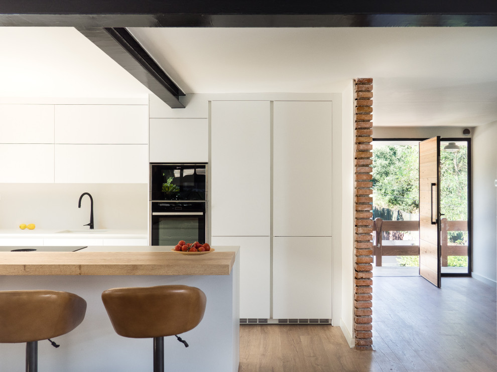 This is an example of a large contemporary single-wall open plan kitchen in Barcelona with an integrated sink, flat-panel cabinets, white cabinets, quartz benchtops, white splashback, engineered quartz splashback, panelled appliances, with island and white benchtop.