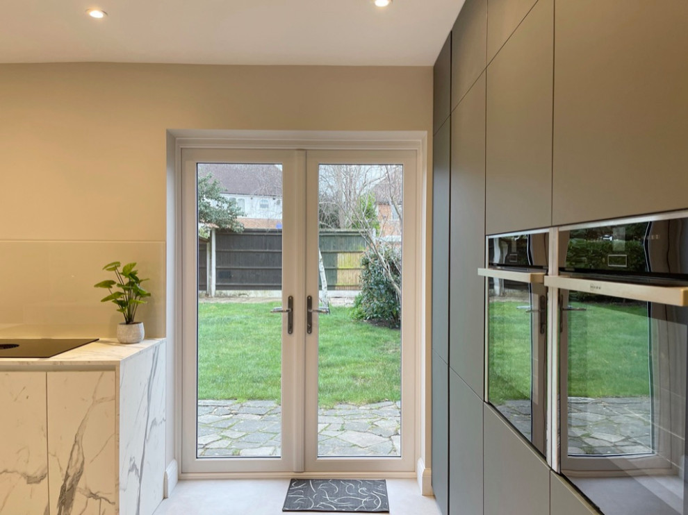 This is an example of a mid-sized modern l-shaped eat-in kitchen in London with an integrated sink, flat-panel cabinets, green cabinets, laminate benchtops, white splashback, glass sheet splashback, black appliances, laminate floors, no island, beige floor and white benchtop.