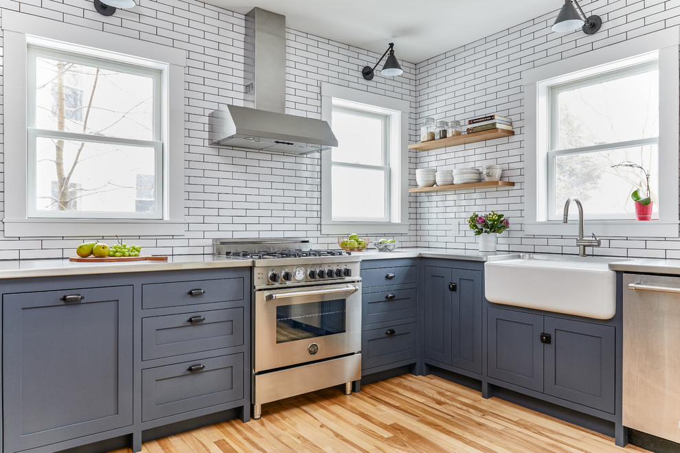 Photo of a transitional l-shaped kitchen in Portland Maine with a farmhouse sink, shaker cabinets, blue cabinets, concrete benchtops, white splashback, stone tile splashback, stainless steel appliances, light hardwood floors, no island and beige floor.