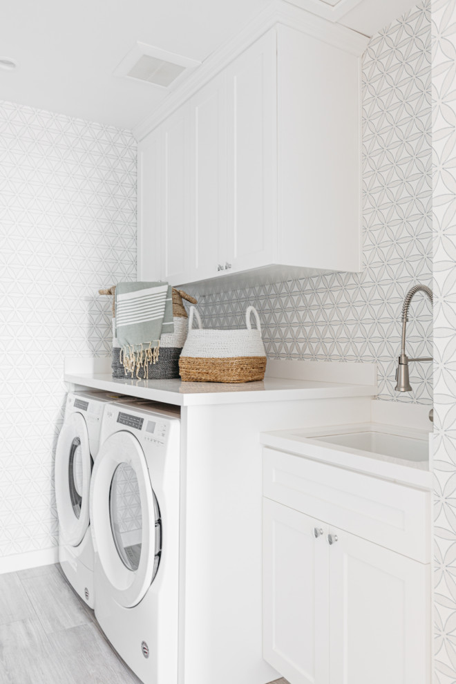Inspiration for a beach style single-wall laundry room in Los Angeles with an undermount sink, shaker cabinets, white cabinets, grey walls, a side-by-side washer and dryer, grey floor, white benchtop and wallpaper.