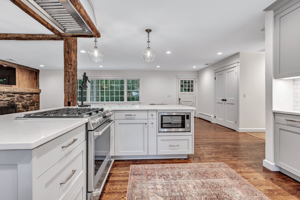 This is an example of a large country u-shaped eat-in kitchen in New York with a farmhouse sink, shaker cabinets, grey cabinets, quartz benchtops, white splashback, subway tile splashback, stainless steel appliances, medium hardwood floors, with island, brown floor, white benchtop and exposed beam.