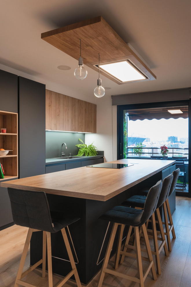 Photo of a large contemporary single-wall open plan kitchen in Valencia with a farmhouse sink, flat-panel cabinets, black cabinets, wood benchtops, black splashback, black appliances, medium hardwood floors, with island and brown benchtop.