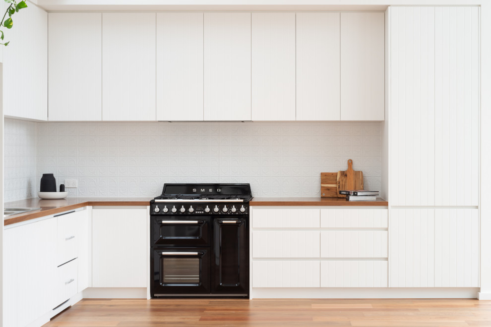 Small scandinavian l-shaped eat-in kitchen in Melbourne with a double-bowl sink, flat-panel cabinets, white cabinets, wood benchtops, white splashback, metal splashback, black appliances, medium hardwood floors, no island, brown floor, brown benchtop and vaulted.