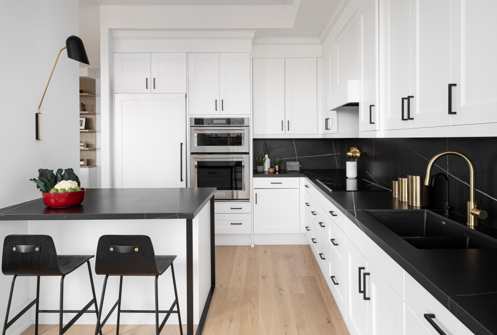 This is an example of a mid-sized scandinavian l-shaped eat-in kitchen in Toronto with an undermount sink, shaker cabinets, white cabinets, black splashback, white appliances, light hardwood floors, a peninsula and black benchtop.