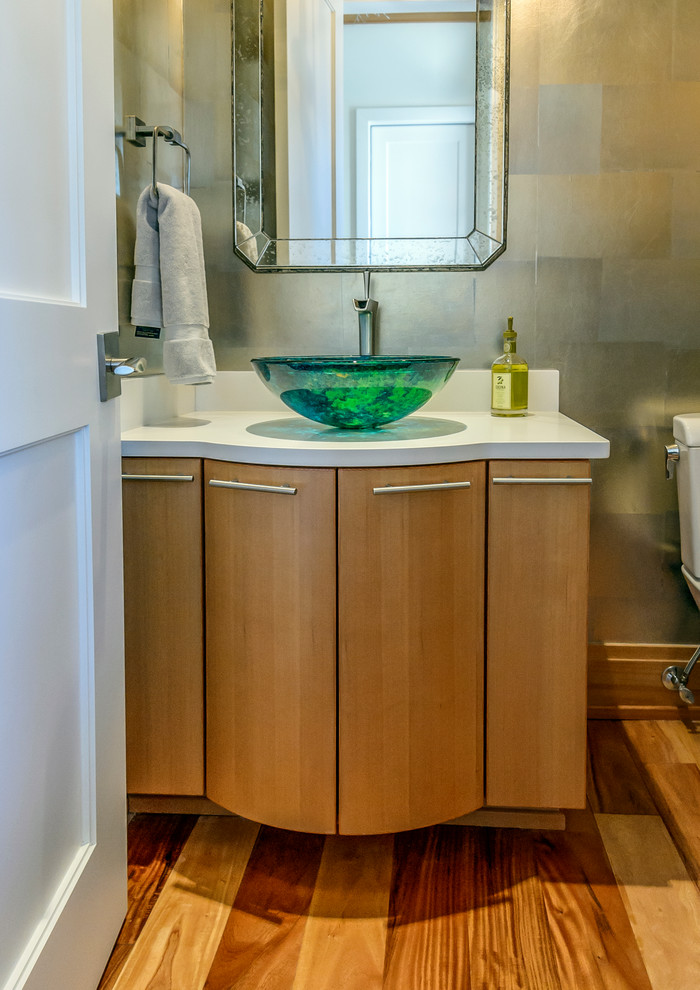 Photo of a small arts and crafts powder room in Charleston with flat-panel cabinets, light wood cabinets, a one-piece toilet, multi-coloured walls, a vessel sink, engineered quartz benchtops, brown floor, medium hardwood floors and white benchtops.