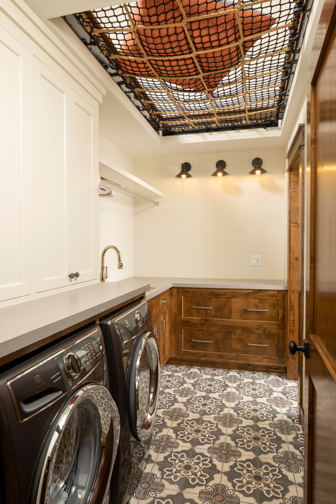 Inredning av en rustik mellanstor l-formad tvättstuga, med en undermonterad diskho, luckor med infälld panel, bruna skåp, laminatbänkskiva, betonggolv, en tvättmaskin och torktumlare bredvid varandra och flerfärgat golv