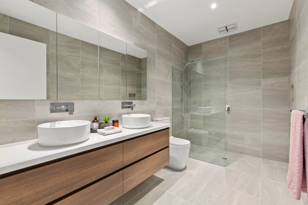 This is an example of a modern 3/4 wet room bathroom in Melbourne with flat-panel cabinets, medium wood cabinets, beige tile, beige walls, a vessel sink, beige floor, an open shower and white benchtops.