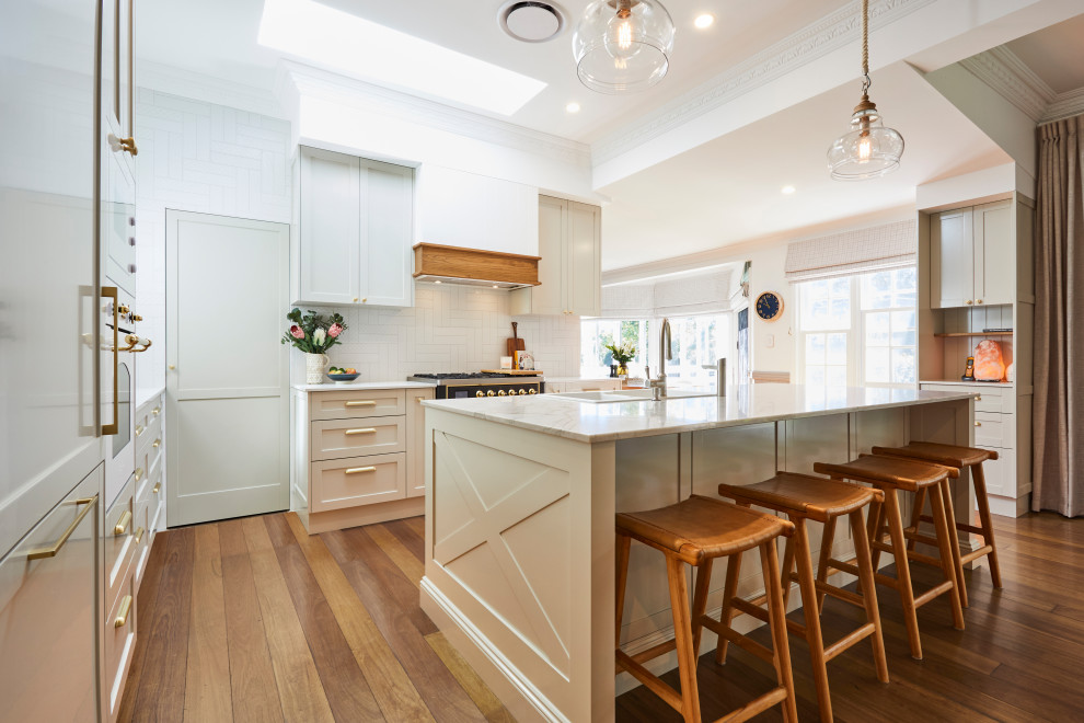 This is an example of a large beach style l-shaped kitchen pantry in Sunshine Coast with an integrated sink, shaker cabinets, white cabinets, quartz benchtops, white splashback, subway tile splashback, black appliances, light hardwood floors, with island, brown floor and beige benchtop.