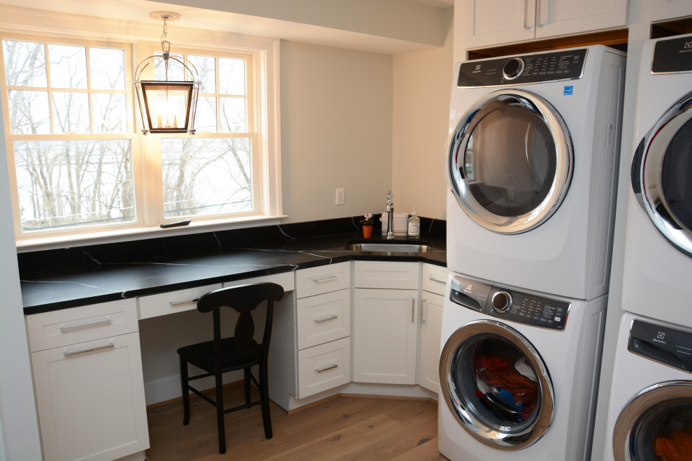 This is an example of a large beach style l-shaped dedicated laundry room in Baltimore with an undermount sink, recessed-panel cabinets, white cabinets, quartz benchtops, white walls, a stacked washer and dryer, brown floor and black benchtop.