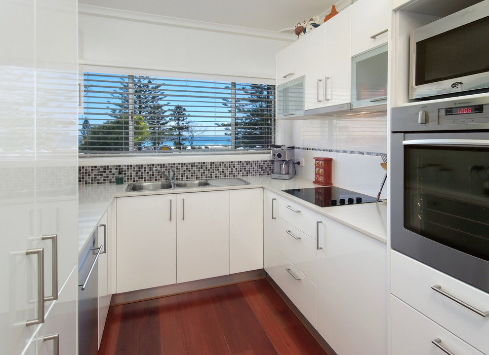 Design ideas for a small contemporary u-shaped separate kitchen in Gold Coast - Tweed with a drop-in sink, flat-panel cabinets, white cabinets, quartz benchtops, white splashback, porcelain splashback, stainless steel appliances, medium hardwood floors and no island.