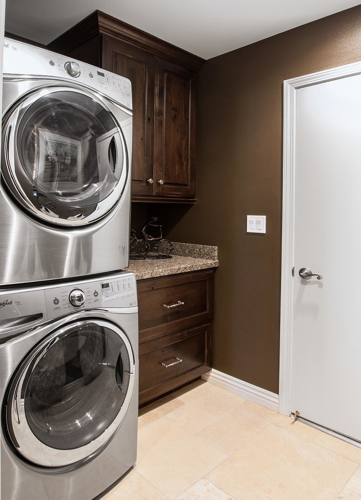 This is an example of a small traditional single-wall dedicated laundry room in Orange County with an undermount sink, recessed-panel cabinets, dark wood cabinets, granite benchtops, brown walls, porcelain floors and a stacked washer and dryer.