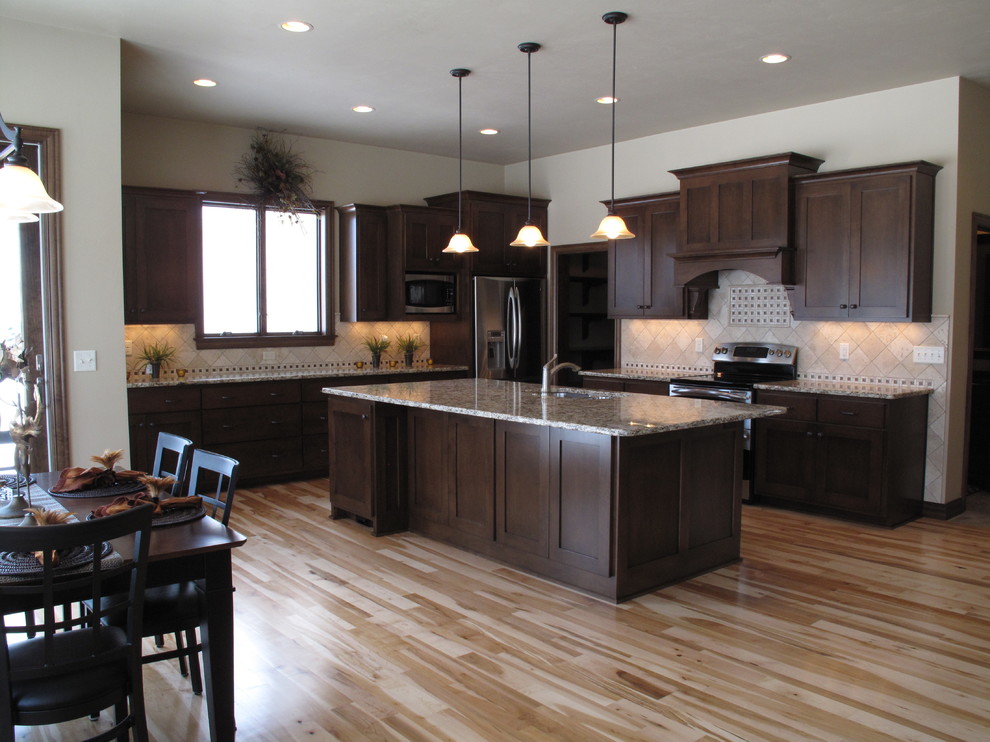 Example of a light wood floor and brown floor kitchen design with an undermount sink, shaker cabinets, dark wood cabinets, granite countertops, stainless steel appliances and an island