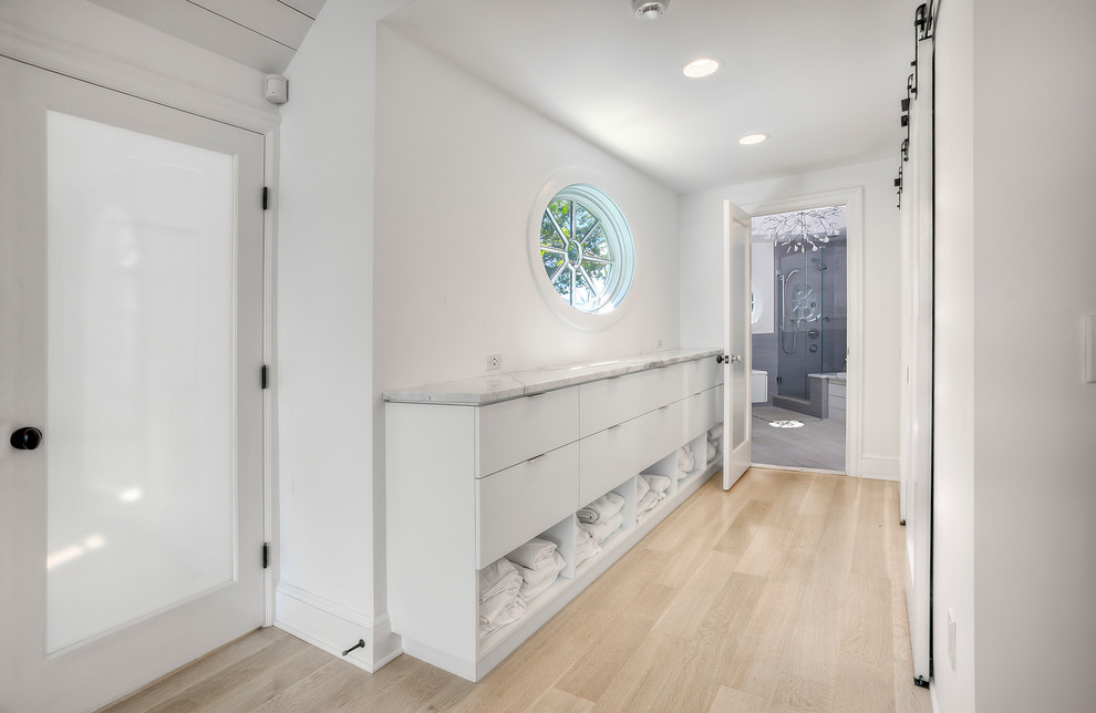Inspiration for a mid-sized beach style master bathroom in New York with flat-panel cabinets, white cabinets, a corner tub, a shower/bathtub combo, a one-piece toilet, white walls, light hardwood floors, a wall-mount sink, marble benchtops, beige floor, a hinged shower door and white benchtops.
