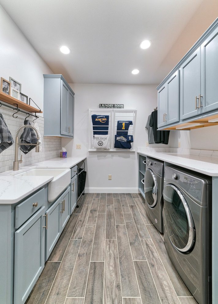 Dedicated laundry room - transitional galley brown floor dedicated laundry room idea in Other with a farmhouse sink, recessed-panel cabinets, blue cabinets, white walls, a side-by-side washer/dryer and white countertops