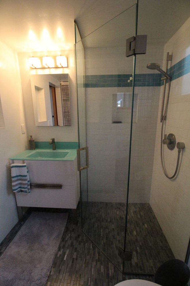 This is an example of a small 3/4 bathroom in San Diego with furniture-like cabinets, white cabinets, a corner shower, a two-piece toilet, white tile, porcelain tile, yellow walls, marble floors, a console sink, glass benchtops, grey floor, a hinged shower door and blue benchtops.