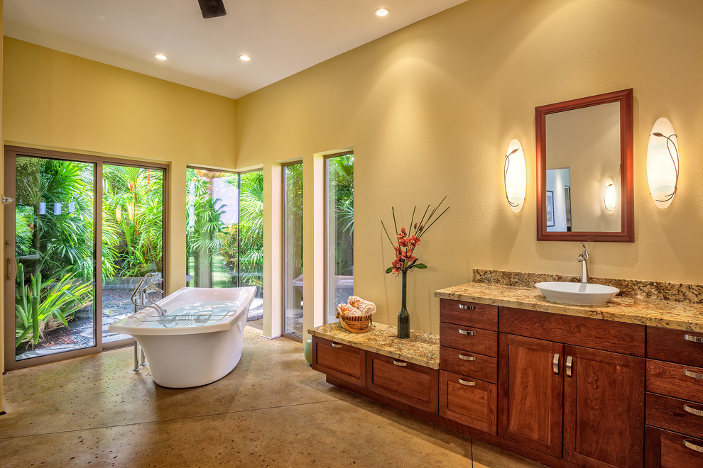 This is an example of an expansive arts and crafts master bathroom in Hawaii with a drop-in sink, recessed-panel cabinets, medium wood cabinets, granite benchtops, a freestanding tub, a curbless shower, a one-piece toilet and concrete floors.