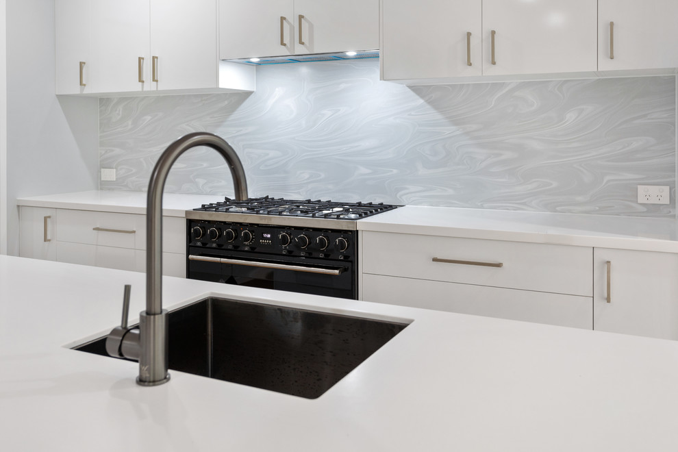 Photo of a mid-sized contemporary galley kitchen pantry in Brisbane with an undermount sink, white cabinets, solid surface benchtops, grey splashback, black appliances, vinyl floors, with island, brown floor and white benchtop.