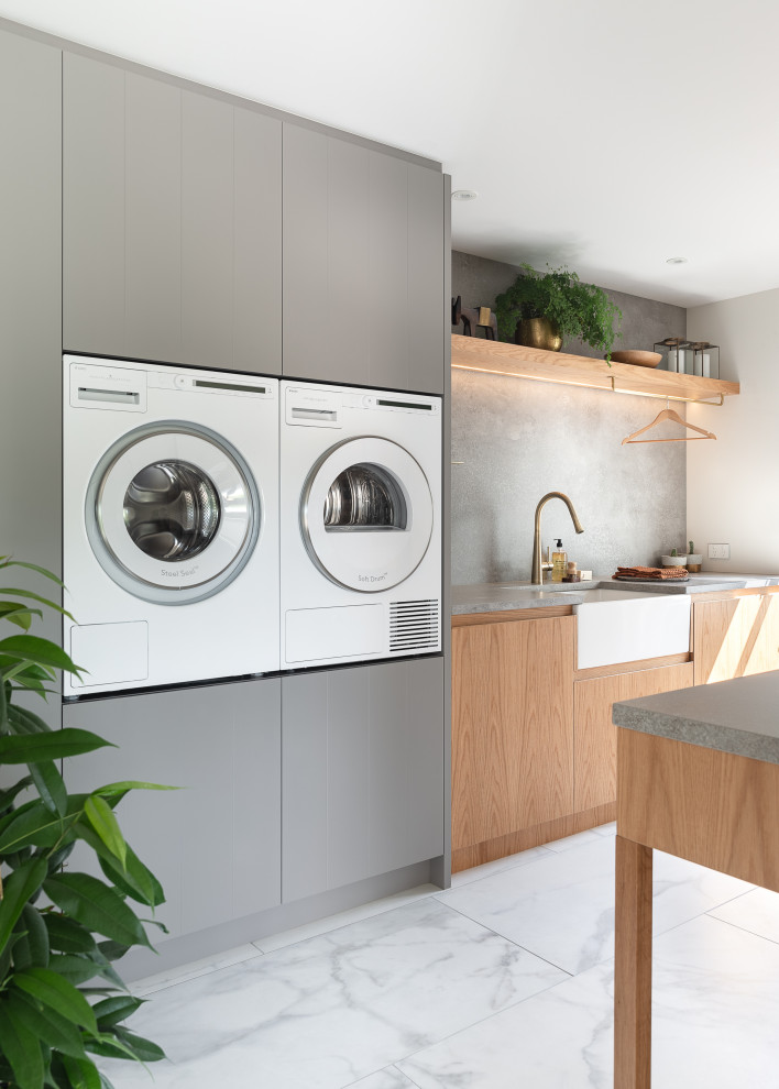 Design ideas for a mid-sized contemporary galley dedicated laundry room in Auckland with a farmhouse sink, flat-panel cabinets, light wood cabinets, quartz benchtops, grey splashback, porcelain splashback, white walls, porcelain floors, a side-by-side washer and dryer, white floor and grey benchtop.