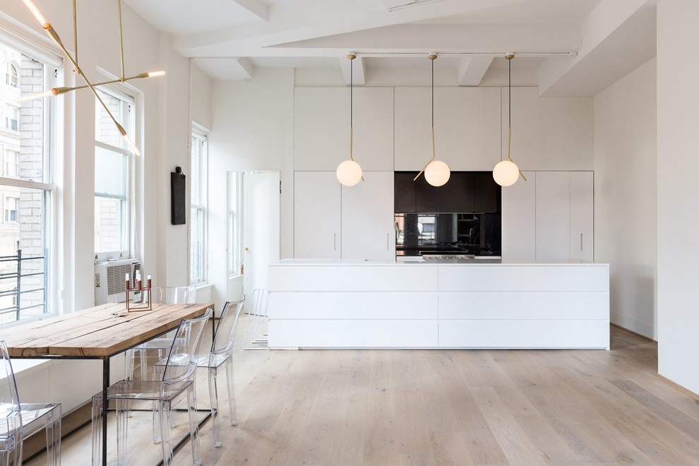 This is an example of a mid-sized scandinavian single-wall eat-in kitchen in New York with flat-panel cabinets, white cabinets, marble benchtops, black splashback, stone slab splashback, black appliances, light hardwood floors and with island.