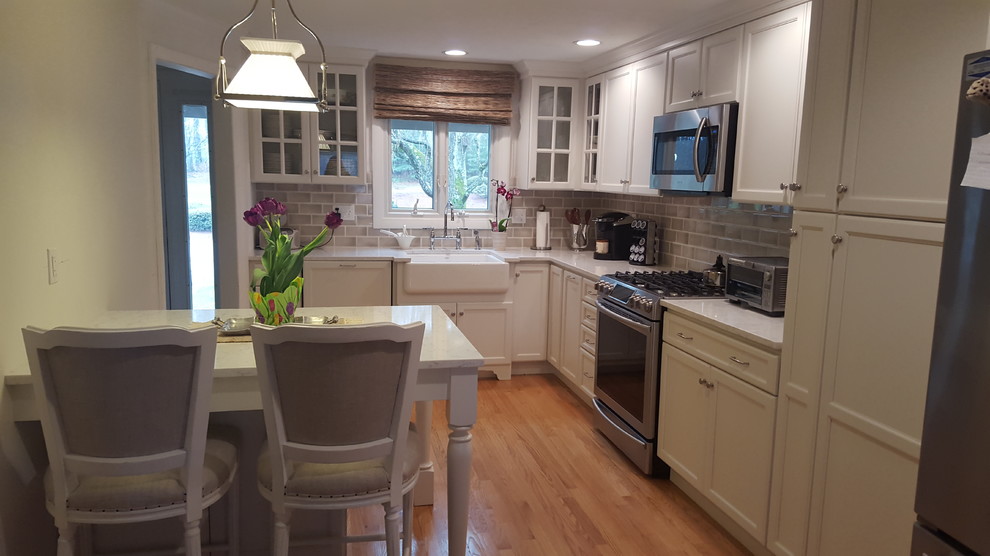This is an example of a small country l-shaped kitchen in Boston with a farmhouse sink, white cabinets, quartz benchtops, beige splashback, ceramic splashback, light hardwood floors and a peninsula.