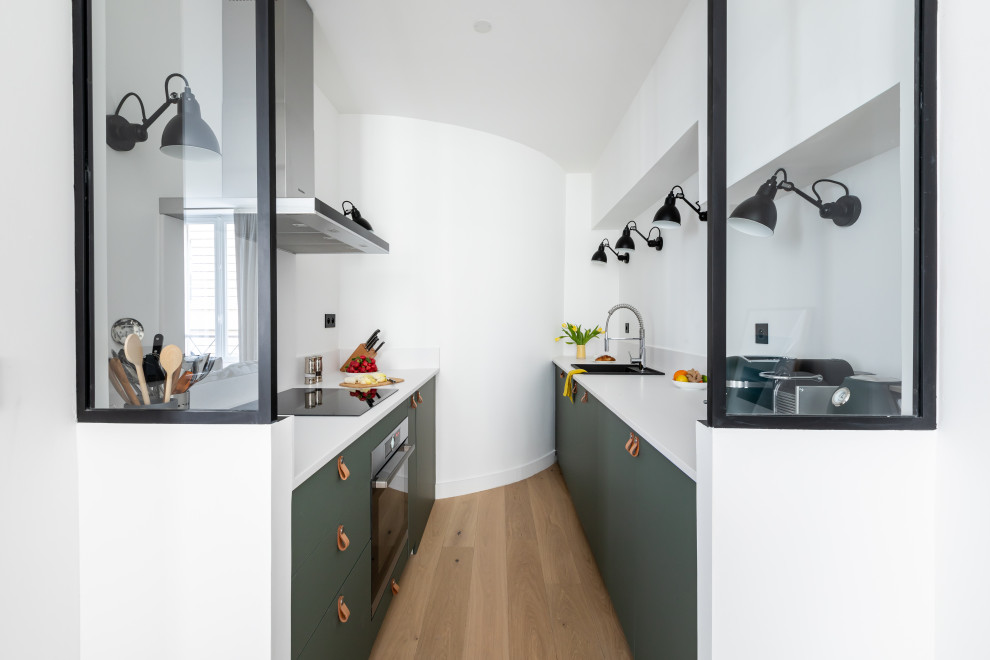 This is an example of a small contemporary galley open plan kitchen in Paris with a single-bowl sink, beaded inset cabinets, green cabinets, solid surface benchtops, white splashback, light hardwood floors and white benchtop.