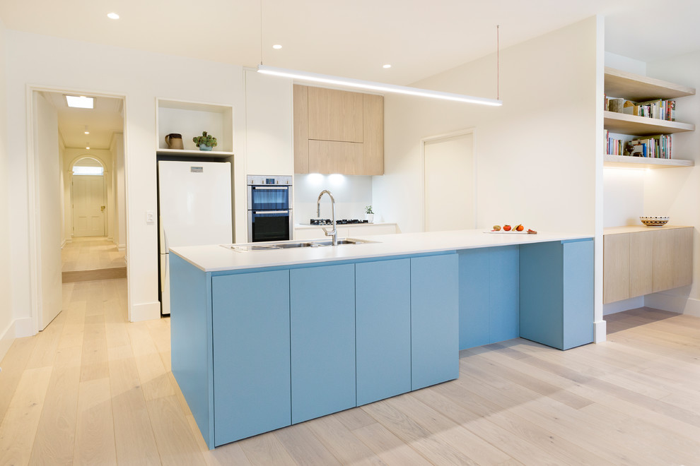 Contemporary galley open plan kitchen in Melbourne with a double-bowl sink, flat-panel cabinets, blue cabinets, wood benchtops, white splashback, white appliances, light hardwood floors, a peninsula and beige floor.