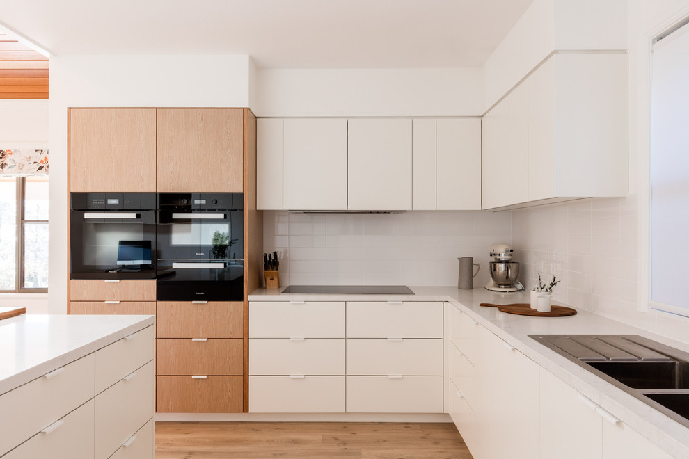 Design ideas for a contemporary l-shaped kitchen in Newcastle - Maitland with a double-bowl sink, flat-panel cabinets, white cabinets, white splashback, subway tile splashback, black appliances, light hardwood floors and with island.