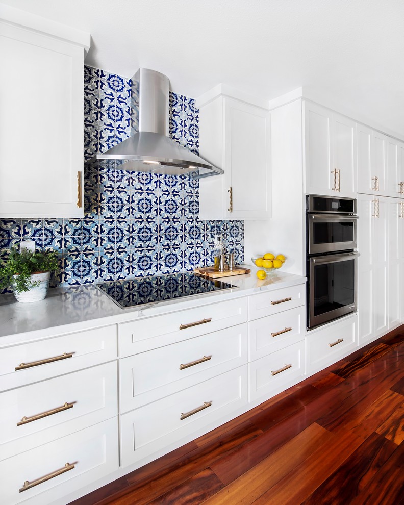 Photo of an eclectic eat-in kitchen with shaker cabinets, white cabinets, quartz benchtops, blue splashback, ceramic splashback, stainless steel appliances, dark hardwood floors, a peninsula and brown floor.