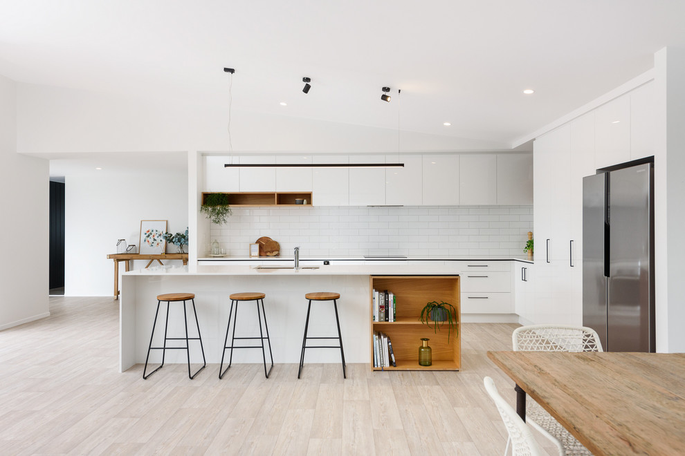 This is an example of a contemporary l-shaped eat-in kitchen in Hamilton with an undermount sink, flat-panel cabinets, white cabinets, white splashback, subway tile splashback, stainless steel appliances, with island, beige floor and white benchtop.