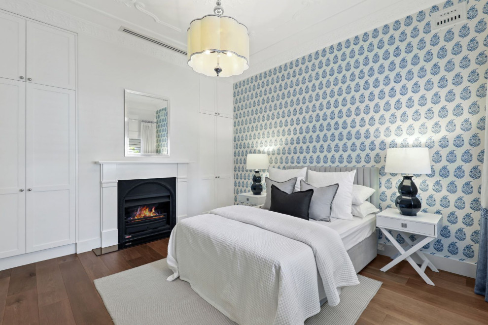 На фото: гостевая спальня (комната для гостей) в стиле неоклассика (современная классика) с белыми стенами, паркетным полом среднего тона, стандартным камином, коричневым полом и обоями на стенах