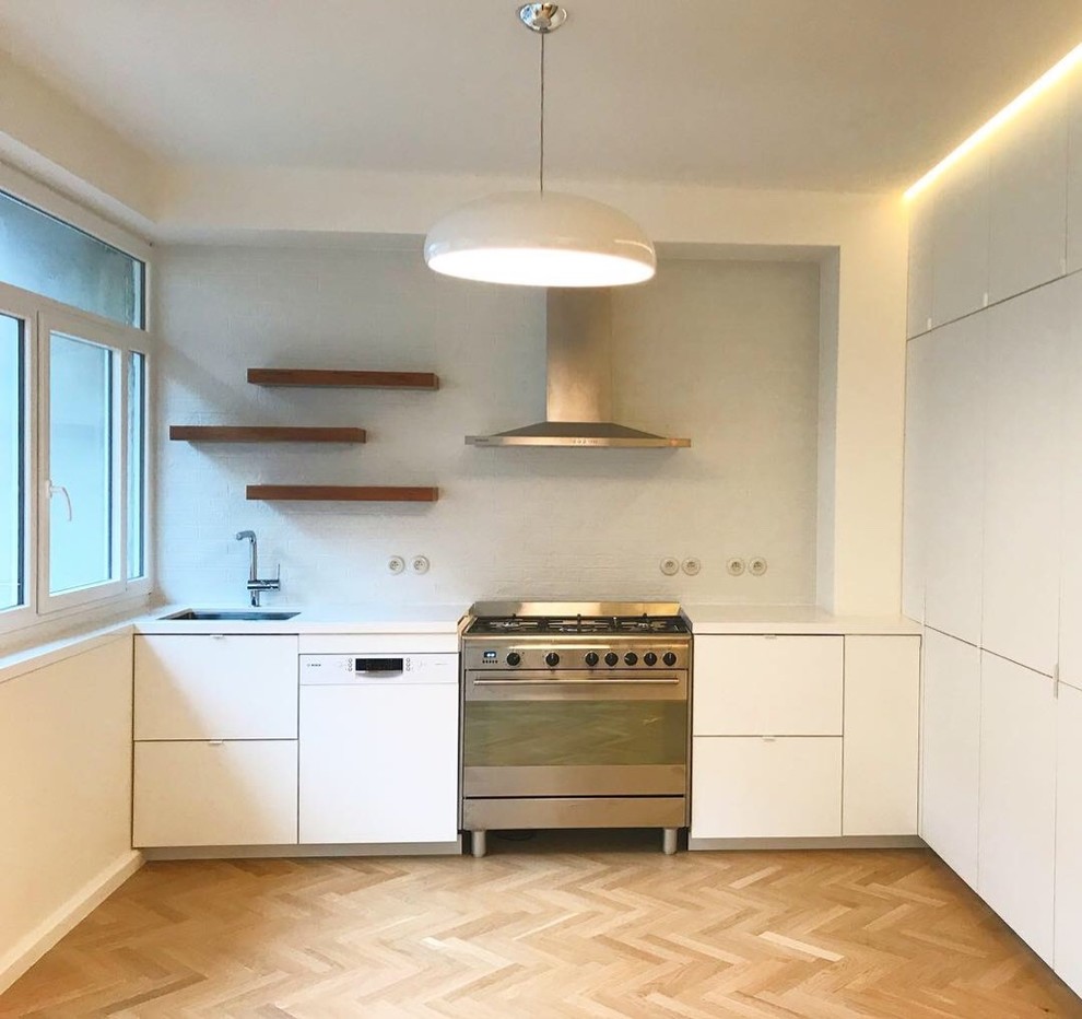 Mid-sized contemporary l-shaped open plan kitchen in Paris with an undermount sink, flat-panel cabinets, white cabinets, granite benchtops, white splashback, brick splashback, stainless steel appliances, light hardwood floors, no island, brown floor and white benchtop.
