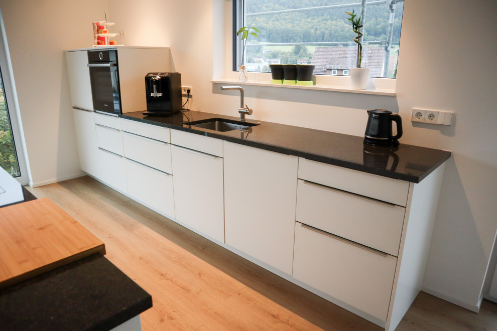 Küche in L-Form mit Einbauwaschbecken, Kassettenfronten, weißen Schränken, Mineralwerkstoff-Arbeitsplatte, Küchenrückwand in Weiß, Küchengeräten aus Edelstahl, Laminat, Kücheninsel, braunem Boden, schwarzer Arbeitsplatte und kleiner Kücheninsel