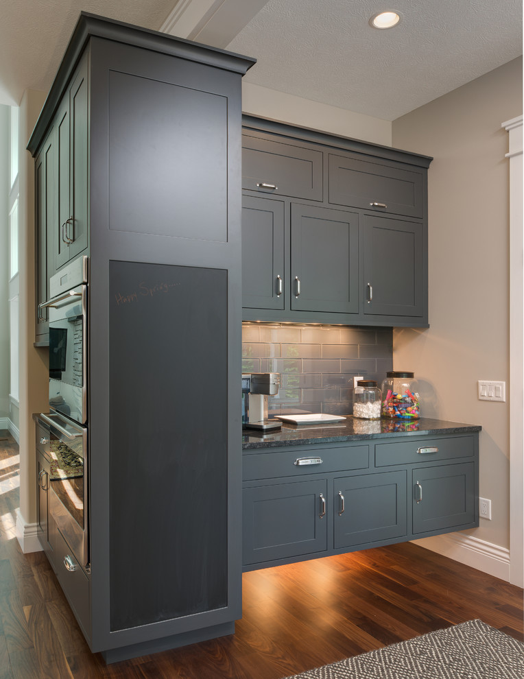 Example of a large transitional single-wall medium tone wood floor kitchen pantry design with an undermount sink, shaker cabinets, gray cabinets, granite countertops, gray backsplash, ceramic backsplash, stainless steel appliances and an island