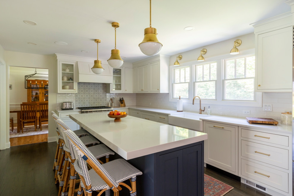 Klassisk inredning av ett stort vit vitt kök, med en rustik diskho, luckor med infälld panel, vita skåp, bänkskiva i kvarts, vitt stänkskydd, stänkskydd i porslinskakel, rostfria vitvaror, mellanmörkt trägolv, en köksö och brunt golv