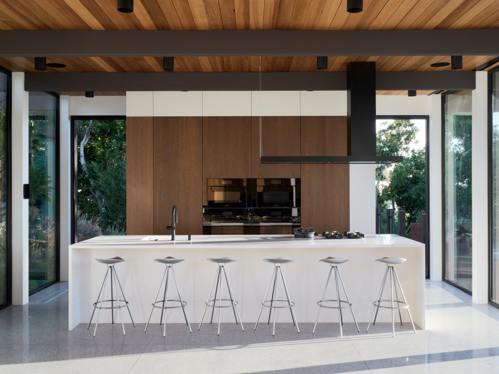 Example of a 1960s galley gray floor, exposed beam and wood ceiling kitchen design in Los Angeles with an integrated sink, flat-panel cabinets, medium tone wood cabinets, black appliances, an island and white countertops