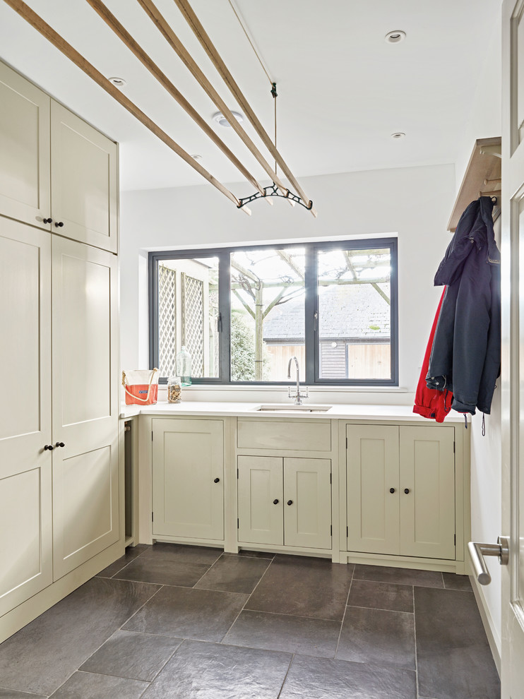 This is an example of a mid-sized beach style galley laundry cupboard in Devon with a drop-in sink, recessed-panel cabinets, light wood cabinets, porcelain floors, a concealed washer and dryer, grey floor, white benchtop, granite benchtops and white walls.