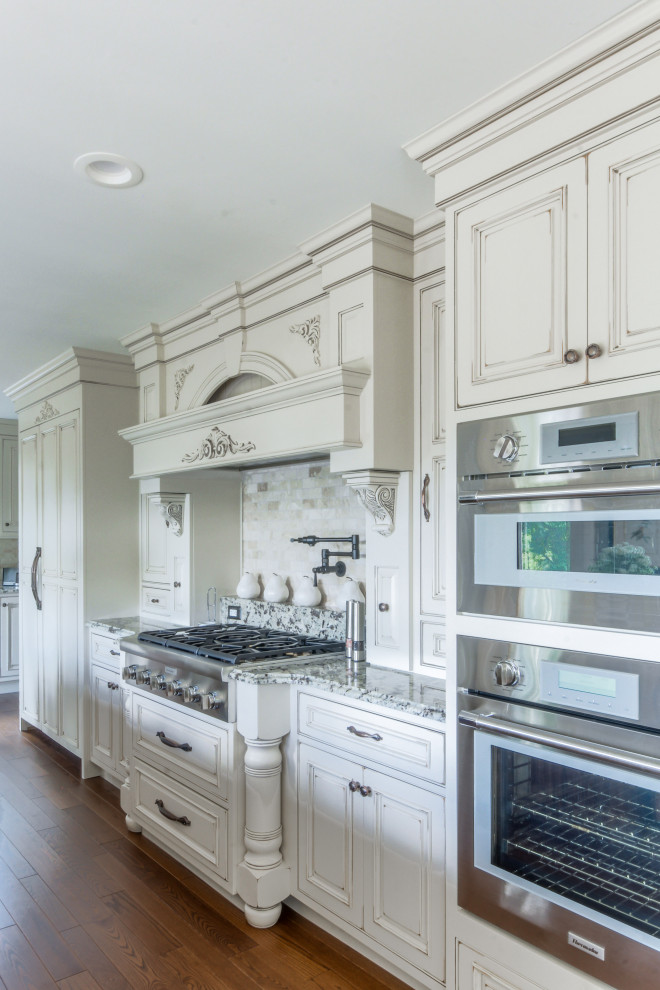 Inspiration for a large traditional l-shaped eat-in kitchen in Detroit with a farmhouse sink, raised-panel cabinets, distressed cabinets, quartz benchtops, beige splashback, marble splashback, stainless steel appliances, medium hardwood floors, with island, brown floor and white benchtop.