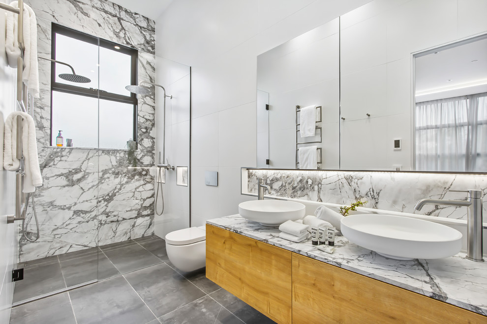 Photo of a contemporary master bathroom in Melbourne with flat-panel cabinets, light wood cabinets, a curbless shower, a wall-mount toilet, white tile, white walls, a vessel sink, grey floor, a hinged shower door and white benchtops.
