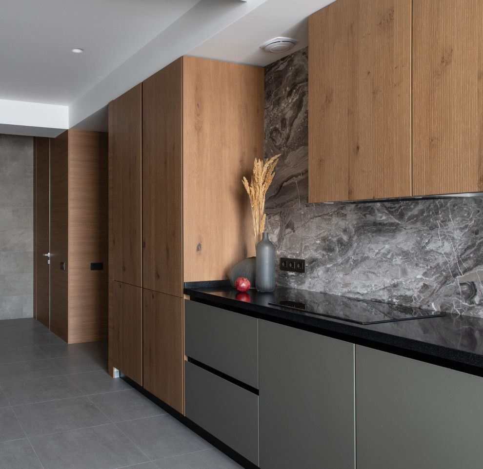 Inspiration for a contemporary single-wall kitchen in Other with flat-panel cabinets, medium wood cabinets, solid surface benchtops, grey splashback, marble splashback, black appliances, porcelain floors, grey floor and black benchtop.