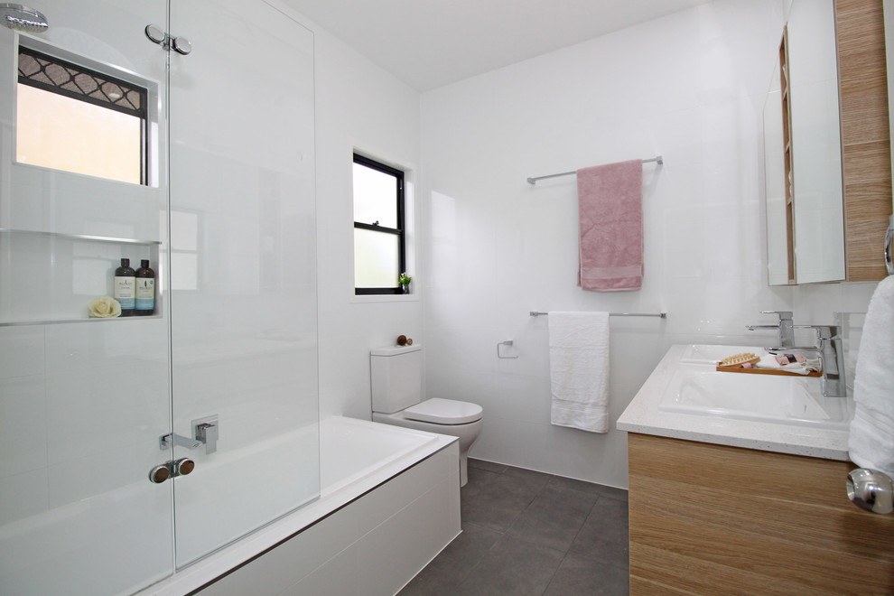 Design ideas for a mid-sized modern kids bathroom in Brisbane with flat-panel cabinets, light wood cabinets, a drop-in tub, a shower/bathtub combo, a two-piece toilet, white tile, ceramic tile, white walls, porcelain floors, a drop-in sink, engineered quartz benchtops, grey floor, a hinged shower door and white benchtops.