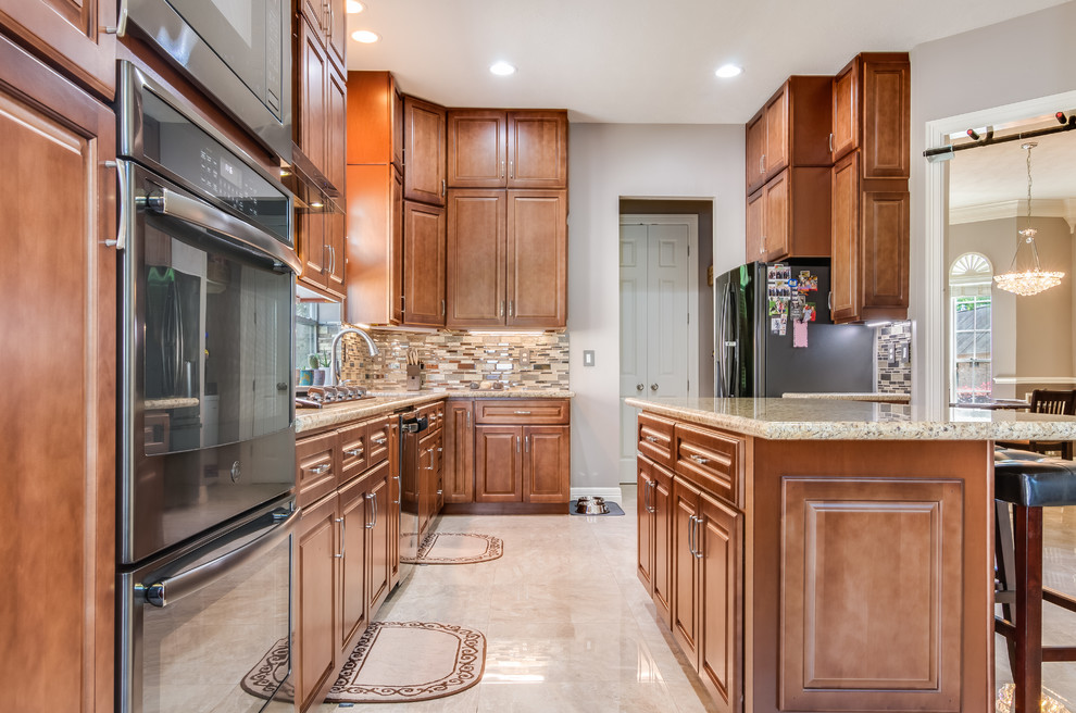 Photo of a large traditional l-shaped eat-in kitchen in Houston with a double-bowl sink, raised-panel cabinets, brown cabinets, granite benchtops, brown splashback, glass sheet splashback, black appliances, marble floors, with island, brown floor and brown benchtop.