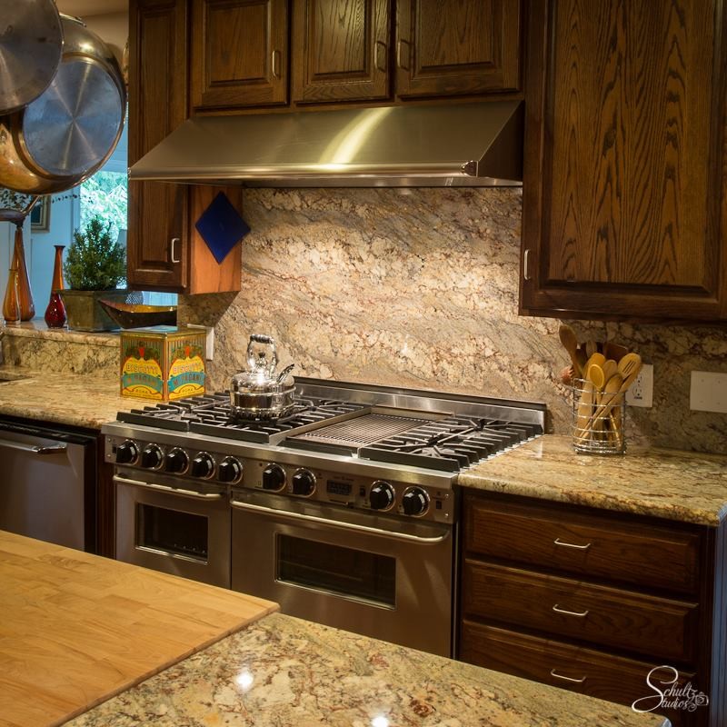 Example of a mid-sized transitional galley enclosed kitchen design with raised-panel cabinets, dark wood cabinets, granite countertops, beige backsplash, stone slab backsplash, stainless steel appliances and an island