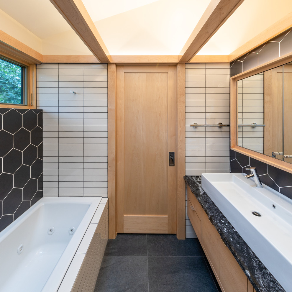 This is an example of a mid-sized modern master bathroom in Cedar Rapids with flat-panel cabinets, light wood cabinets, a drop-in tub, an alcove shower, a one-piece toilet, white tile, ceramic tile, white walls, ceramic floors, a vessel sink, granite benchtops, black floor, an open shower and black benchtops.