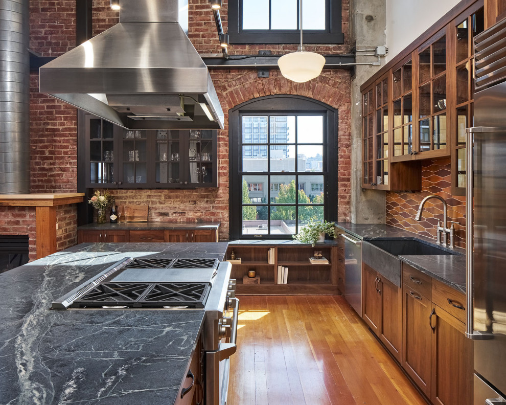 Mid-sized industrial l-shaped open plan kitchen in Portland with a farmhouse sink, glass-front cabinets, dark wood cabinets, soapstone benchtops, multi-coloured splashback, ceramic splashback, stainless steel appliances, medium hardwood floors, with island, brown floor, black benchtop and exposed beam.