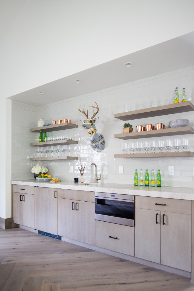 Photo of a large country single-wall wet bar with a drop-in sink, flat-panel cabinets, medium wood cabinets, quartzite benchtops, white splashback, porcelain splashback, light hardwood floors, beige benchtop and beige floor.