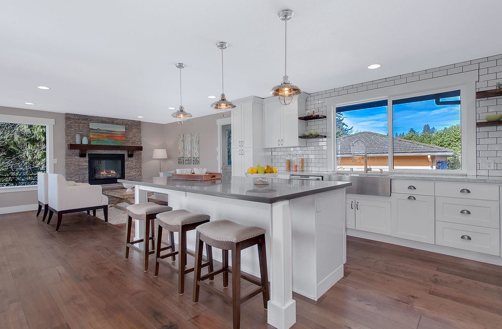 Inspiration for a mid-sized transitional l-shaped open plan kitchen in Seattle with a farmhouse sink, shaker cabinets, white cabinets, white splashback, subway tile splashback, stainless steel appliances, dark hardwood floors, with island, quartz benchtops and brown floor.